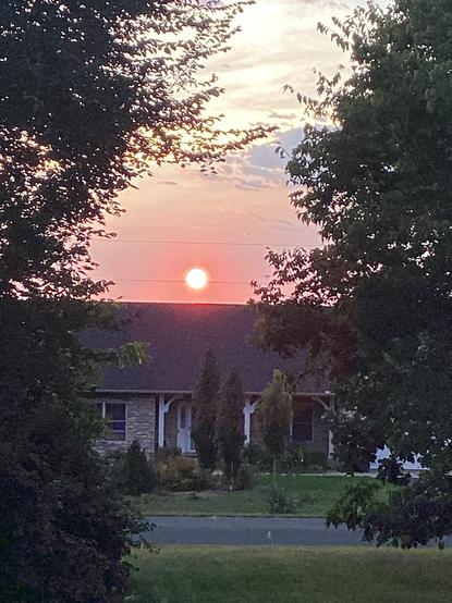 A sunset view of the sun just above the roof of a house,. The view is framed by large trees on both sides. The sunset is unusually dim and clear due to smoke from wildfires that cause the light to be a very unusual shade of orange.