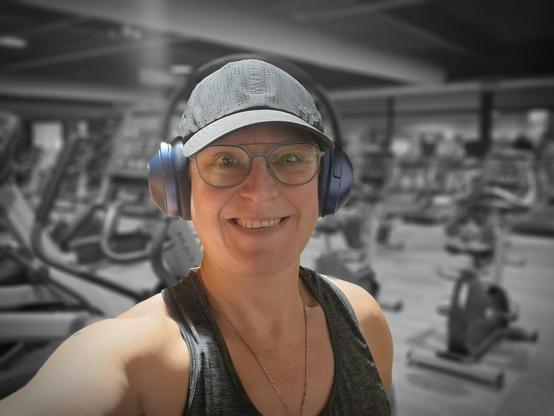 Selfie of a person in the gym. Smiling at the camera. Used a filter to have me in color and the background is black and white. Wearing a grey top, black cap, blue headphones and blue-rimmed glasses. In the background are several cardio machines.