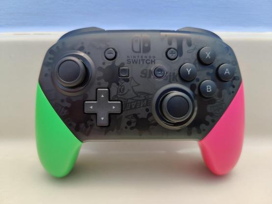 A Nintendo Switch Pro controller of the Splatoon 2 design with neon pink and green handles, and dark gray art on the face of different ink splatter and some graffiti from the game like the cartoon squid with Splatoon language text underneath it.