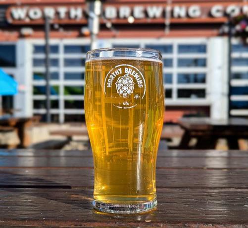 A pint of Adult Lemonade Radler from Worthy Brewing of Bend, Oregon. The logo on the pint glass is well worn and slightly rubbed off. The beer in the glass is a brassy yellow color. The glass sits on a picnic table in the brewery's beer garden, and you can see a blurred background with the brewery's name in large letters on the side of the building.