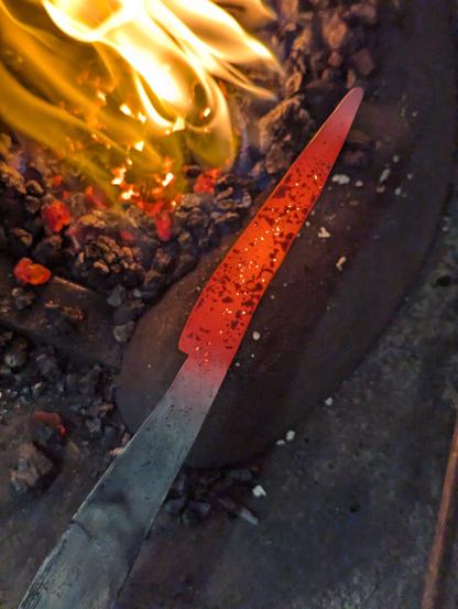 Vue plongeante sur le foyer de forge , sur le côté une lame de couteau de table forgée rougeoyante, sortie du feu.
