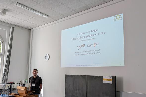 Foto vom Vortragendenbereich des Workshops. Christoph Broschinski steht am Sprechenden-Pult, im Hintergrund ist die Startfolie zu sehen.