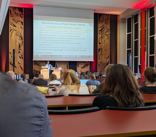 Foto aus dem Zuschauendenraum auf die Bühne. Am Sprechenden-Pult steht der Vortragende, im Hintergrund seine Folien, dazwischen Hinterköpfe von Zuschauenden.