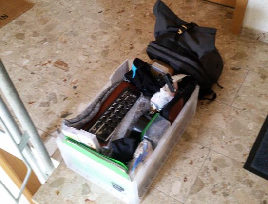 In einem gefliesten Treppenhaus steht eine 42l Eurobos vollgepackt mit schwer zu erkennenden Sachen, daneben lehnt eine schwarze Fahrradtasche. Wenn man es weiß, ist in der Box zu sehen: ein mobiler Gitarren-/Mikrophonverstärker, eine Plastedose die einen in Socken eingepackten Field-Recoder enthält, eine kleine Laptop-Hülle, in der die Mikrophonkapseln und die Stereoschiene sind, Beutel und Taschen mit Leinen und Zubehör, eine Dose mit einem leckeren Brot mit frischen Sprossen.