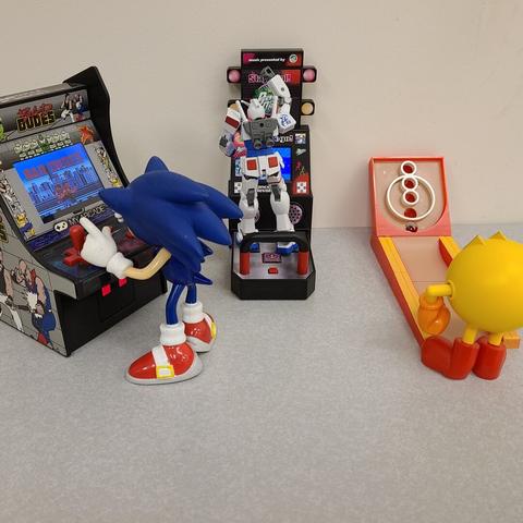 Toy photograph. In the photo, there are three arcade setups, from left to right, Sonic the Hedgehog is playing a miniature Bad Dudes arcade machine; a Gundam unit is playing Dance Dance Revolution in the middle, and to the right Pac-Man is playing skee-ball.