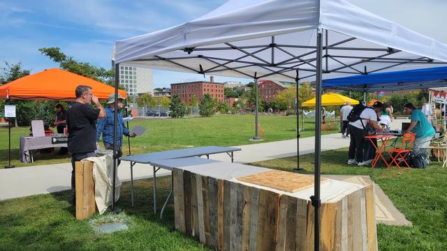 A fifteen-foot-wide tented pop-up shelter on a grass field. Two light-skinned men stand behind the tent, looking as thought they are surveying the space, drinking coffee, or both. There are two tables partially wrapped in light wood planking. In the distance are a bright orange tent, a bright yellow tent, and a royal blue tent with several people gathered around orang folding chairs and tables.