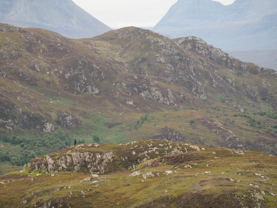 low rocky hills, mountains behind, tiny solitary figure among to centre left
