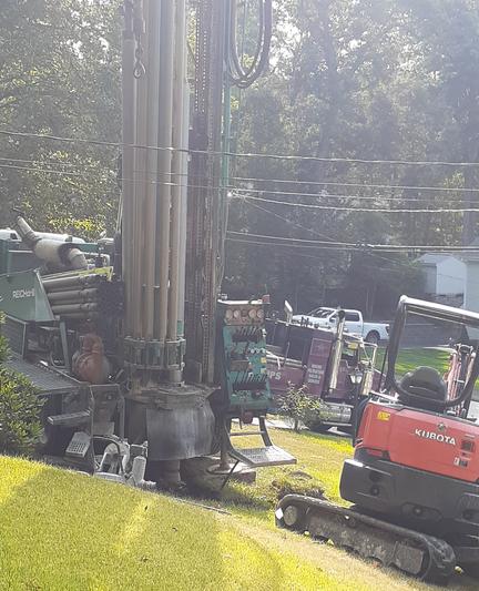 Photo of a front yard with two large construction vehicles on the lawn, and an additional truck on the driveway in the background. The vehicle on the left has a very tall vertical component that is used to bore a hole into the ground.