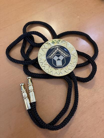 A round gold and black pendant is displayed, featuring a geometric design and an emblem of a house at its center. It is attached to a black braided cord with golden ends, resting on a wooden surface.