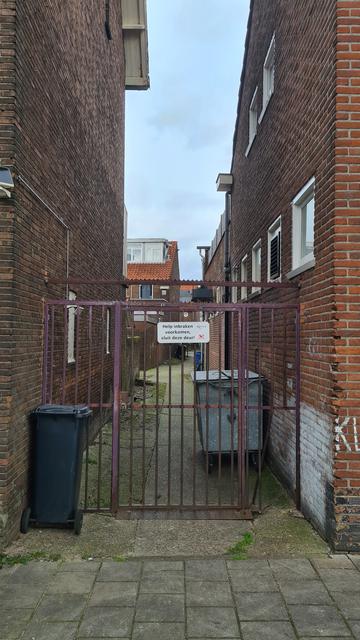 Rusted iron gate between two red brick buildings closing off the entrance to a backalley, with a large hole in the lower right corner because some of its vertical bars are missing and a sign on it reading: help prevent burglaries, close this door.