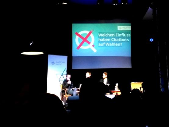Ein Podium, hinten auf der Leinwand der Titel: "Welchen Einfluss haben Chatbots auf Wahlen?"