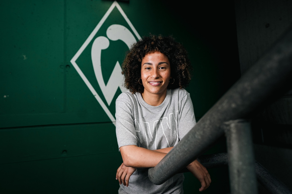 Pressefoto von Amira Arfaoui, die vor einer Werder-Raute im Weserstadion in die Kamera lächelt.