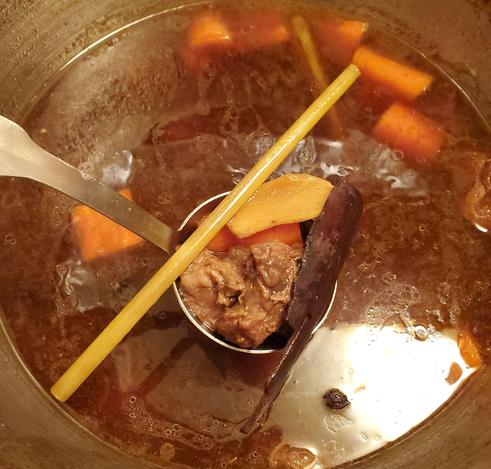 Vietnamese beef & carrot stew cooking in large pot. A ladle is raised in pot. In the ladle is beef, carrot, ginger, lemongrass stick & Chinese cinnamon stick aka cassia bark.
