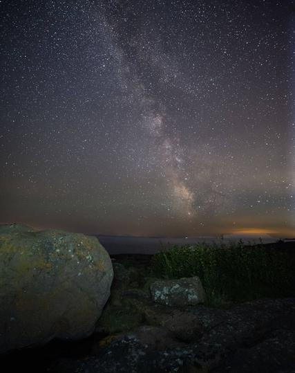 A photograph of the Milky Way over Lake Superior. There is a large boulder and some plants in the foreground.
