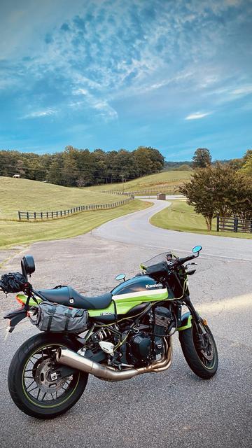An idyllic photograph of Bayport's Kawasaki Z900rs motorcycle parked along the side of a twisty curvy road nestled among grassy green hills with picket fences. Beyond the hills a forested woods can be seen along with wispy clouds among the blue sky.