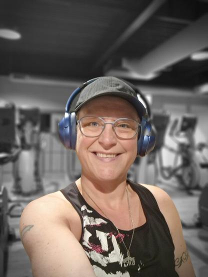 Selfie of a person in the gym. Smiling at the camera. Used a filter to have me in color and the background is black and white. Wearing a black top with white accents, black cap, blue headphones and blue-rimmed glasses.