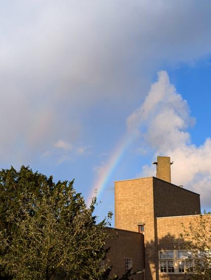Rainbow over a school building with trees in the foreground and white fluffy clouds behind.