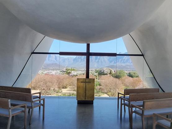View across  the minimalist lectern of a church. The plain window is framed by curved white concrete which looks like drapery, and givesa a panoramicvVista of the mountain range behind it.