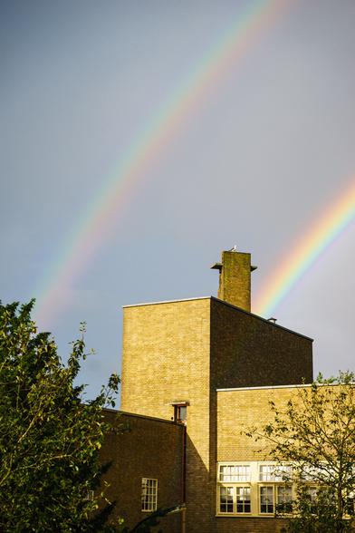 Double rainbow over a school building with trees in the foreground and a gray cloud cover behind.