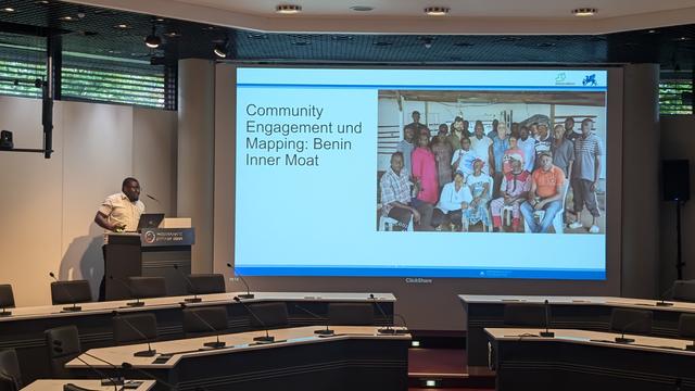 Photo showing a lecture hall. A man is standing at the podium, a large screen shows a slide with a group photo and the title "Community Engagement and Mapping: Benin Inner Moat".