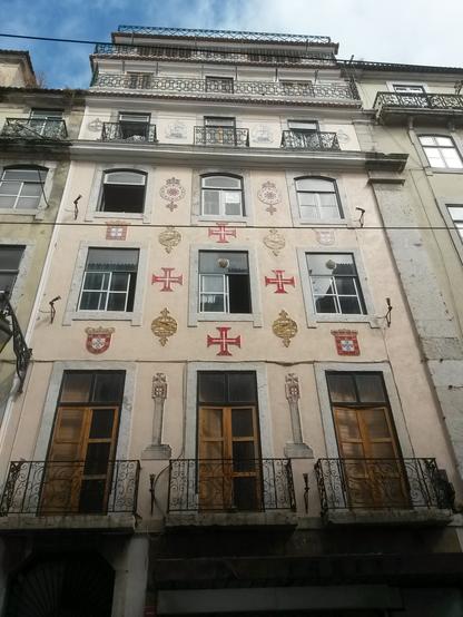 A six store XVIIIth century austere building, painted light pink with 3 stone framed glass windows, mostly square, rows in Lisboa's 'Baixa'. Those on the first floor are wooden door-windows. Between the windows several tile medallions with 'manuelino" late gothic inspired motifs : the Order of Christ cross, national  portuguese coats of arms, armillary spheres, caravels and cross topped marker stones [that used to be left by the portuguese sailors along the african west coast as they proceeded their sailing exploration to the south]. The building's resident families have been (illegally) evicted, with police support, during the pandemic (2020, September) by the owner to give way to short term rentals