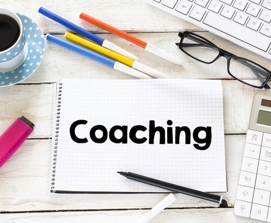Notepad on a desk with the word 'coaching'. There are other stationery items and a cup of black coffee.