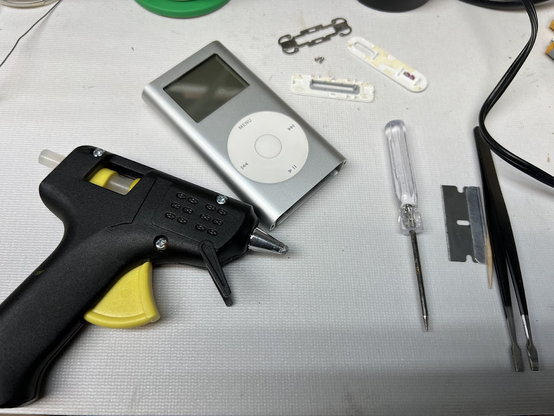 A silver iPod Mini (second generation) with the plastic caps and retaining clips removed, sitting on a workbench with several small tools, including a very cheap-looking hot glue gun.