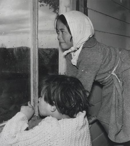 The image features a vintage photograph of two young children peering out of a window. The child in the foreground is wearing a white sweater and has their hands on the sill, while the other child is slightly behind them. They appear to be observing something outside, possibly the rain mentioned, as they are looking down from an upper floor. The lighting suggests it might be early morning or late afternoon due to the soft glow. There are no visible texts in the image.