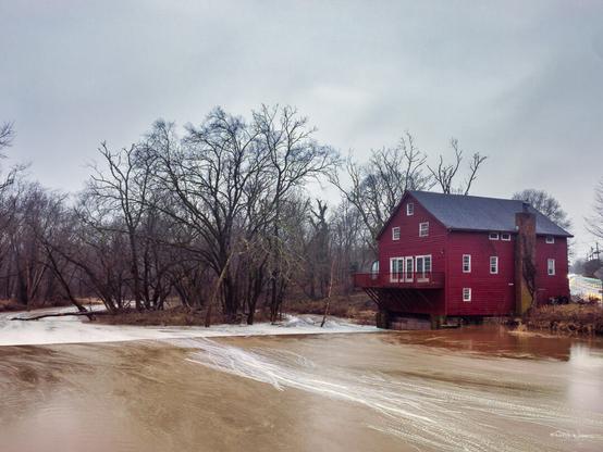 The photograph showcases Opie's Grist Mill, a historic structure perched by the side of Dead Tree Run Bridge in the Bridgepoint Historic District of Montgomery Township, Somerset County, New Jersey. The mill, clad in red panelling, stands out against a winter backdrop with its vibrant colour. It is a multi-story building featuring a prominent balcony overlooking the river. The foreground is dominated by the muddy waters of The Rock Brook, displaying a smooth texture with ripples and reflections that suggest a rapid flow. The bare trees surrounding the mill create a stark, almost solemn atmosphere. The sky is overcast, casting a diffuse light that softens the entire scene, enhancing the mill's prominence. This tranquil setting captures the essence of historic rural America, where such mills were once the heart of local communities.