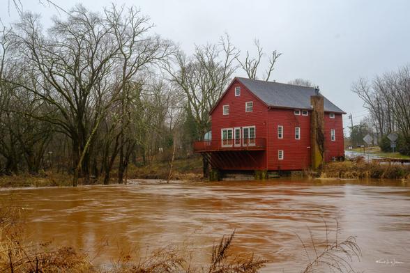 The photograph showcases Opie's Grist Mill, a historic structure perched by the side of Dead Tree Run Bridge in the Bridgepoint Historic District of Montgomery Township, Somerset County, New Jersey. The mill, clad in red panelling, stands out against a winter backdrop with its vibrant colour. It is a multi-story building featuring a prominent balcony overlooking the river. The foreground is dominated by the muddy waters of The Rock Brook, displaying a smooth texture with ripples and reflections that suggest a rapid flow. The bare trees surrounding the mill create a stark, almost solemn atmosphere. The sky is overcast, casting a diffuse light that softens the entire scene, enhancing the mill's prominence. This tranquil setting captures the essence of historic rural America, where such mills were once the heart of local communities.