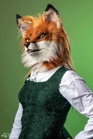 Bust portrait photo of a realistic vixen fursuit. She is wearing a white shirt and green dress in front of a lighter green background. Her golden hair is a bit out of place.