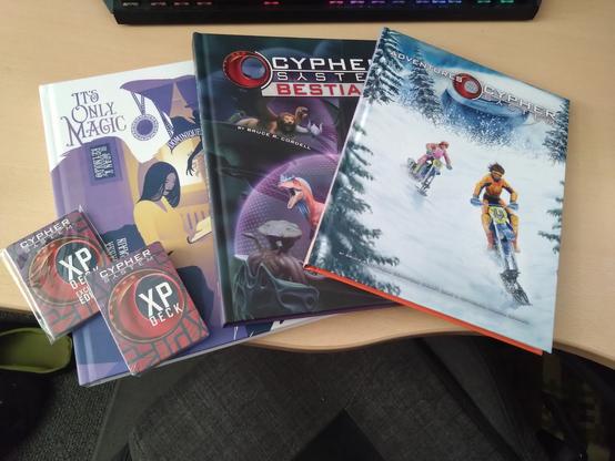 A collection of books laid out on a table. 
Left to right: "It's Only Magic" , "Cypher System Bestiary" and "Adventures in the Cypher System" along with two packs of foil XP cards to be used with the game.