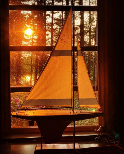 Color image of a 1920s toy pond yacht. Image shows the sun setting over a lake through a stand of tall pine trees, a red and black hulled pond yacht in its cradle, sails up, waiting for its next adventure.