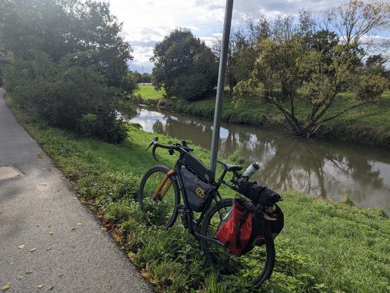 Ein mit Rahmentaschen und Gepäckträger bepacktes Gravelbike, das am Niddaufer an einem Masten angelehnt wurde.