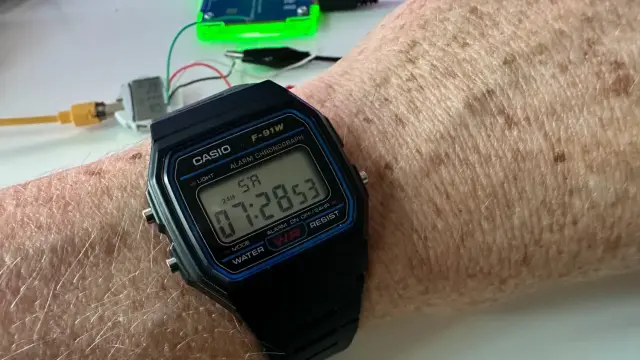 Casio F-91W wrist watch showing "SR" for Sunrise at 07:28:53 with a disassembled watch in the background