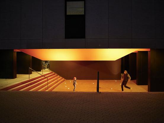 Beneath a modern structure, a child and an adult move in opposite directions under the soft but vivid lights of the night. The geometric shapes of the building frame their interaction, giving the scene a sense of balance between architectural precision and human spontaneity.