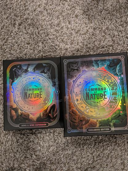 Two black boxes containing the deck building card game Command of Nature. The boxes have shimmering designs that make it look like the logo is crackling with magical energy. 

The background is a carpeted floor because we have no table space in this house, every flat surface is dedicated to toy display
