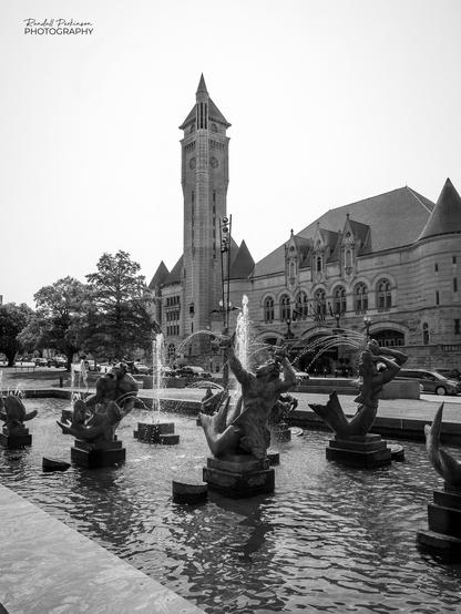 Large stone train station building with a tall tower behind a fountain with multiple statue figures, many of which are shooting water as part of the fountain.