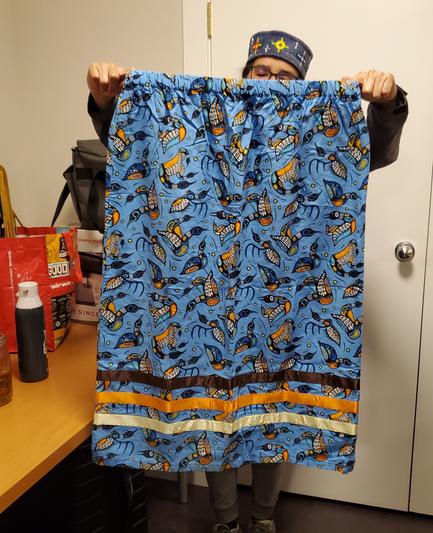 An Indigenous Auntie, holding up her blue loons art fabric ribbon skirt. Brown, orange & gold ribbons at bottom of skirt.