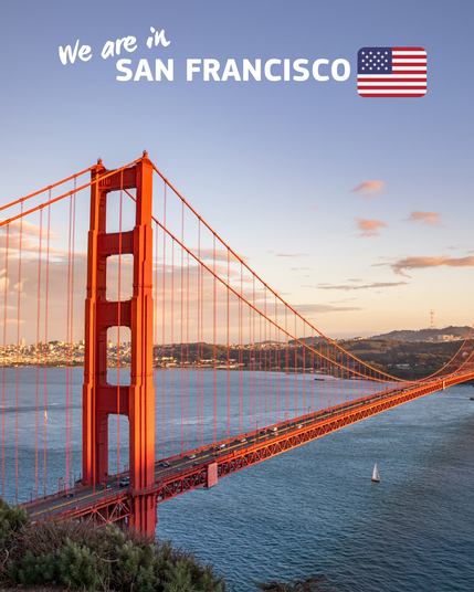A scenic view of the Golden Gate Bridge in San Francisco during sunset, with the bridge's iconic red structure prominently stretching over the water. The sky is clear with a few clouds, and the cityscape of San Francisco can be seen in the background. The text 'We are in San Francisco' is displayed at the top along with a small American flag
