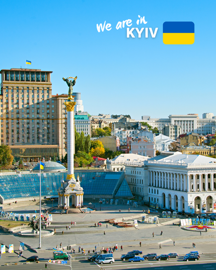 A vibrant view of Independence Square (Maidan Nezalezhnosti) in Kyiv, Ukraine, featuring the Independence Monument, a tall white column with a golden statue on top. Surrounding the square are various buildings, including a large hotel and other architectural landmarks. The Ukrainian flag is visible, and the text 'We are in Kyiv' is displayed at the top with the Ukrainian flag beside it.