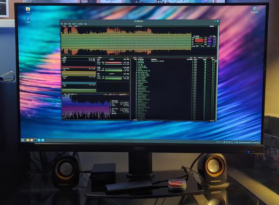A large computer display sitting on a desk with its new stand. It's powered on, with the Linux utility btop running in a shell and colorful wallpaper behind the window.

A pair of cheap speakers are on either side of the stand, and various objects are sitting on top of the tempered glass base.