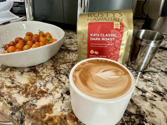 A small white mug filled with a frothy cappuccino sits in front of a bag of Rusty’s Hawaiian Ka’u Classic Dark Roast coffee beans.