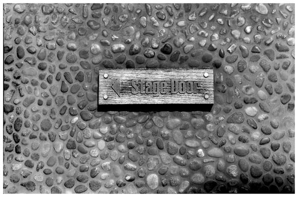 A grainy black and white image of a sign that reads, 'Stage Door', fixed on to a pebble stone wall.