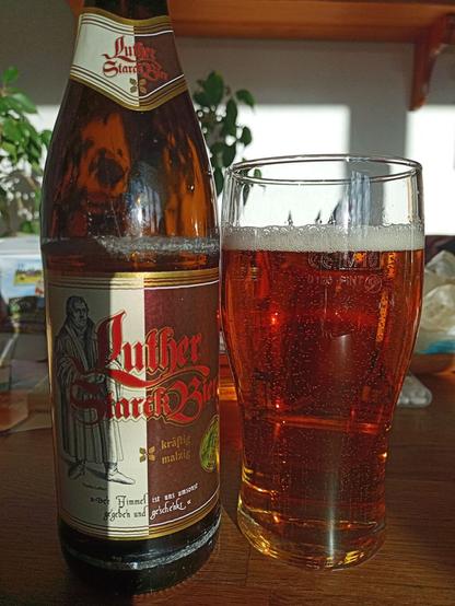 Auf einem.Tisch stehen ein Bierglas, das mit hellbraunen klaren Bier gefüllt ist und daneben die Bierflasche