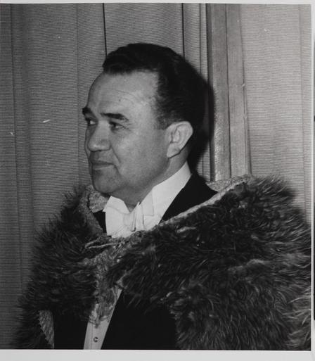 The image is a vintage photograph of a man, likely a well-known opera singer named Inia Te Wiata. He is standing in front of a curtain and wearing a suit with a fur lining or cloak draped over his shoulders. The man is facing to the side, presenting a profile view. There is no visible text in the image.