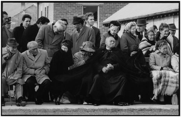 A black and white photo of a group of people at a funeral. They are dressed in attire typical for the 1970s, with some wearing ties visible on men's outfits. The attendees exhibit various emotions, with some bowing their heads in respect, while others maintain more upright postures. They are gathered outside a building, likely a church given the context of a funeral. There is no text visible in the image.