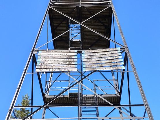 Tekst: "Velkommen til Mistberget
Toppen er nådd
Du står nå oppe i 663moh
Går du opp 16m til er du oppe i tårnhytta

Det første tårnet av tømmer ble bygd 1928
Tårnet som står nå i dag ble bygd i 1938
Takk for besøket
God tur hjem!"