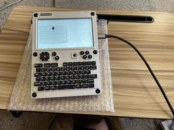 The uconsole is completed and powered on. The Raspberry OS welcome screen shows in a small 720p display above a squishy rubber keyboard. The unit is connected to power with a USB-C.