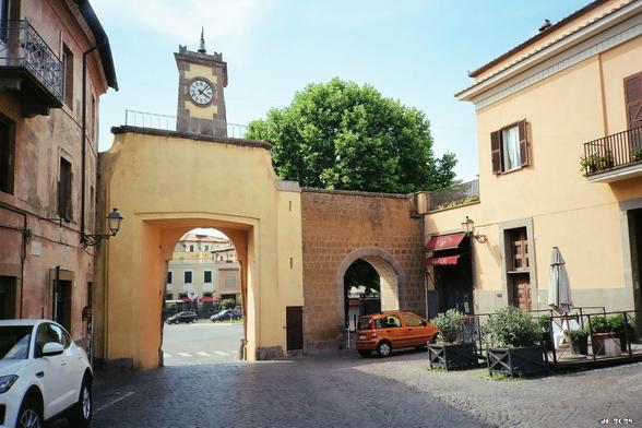 Farbfoto aus der kleinen historischen Stadt Tuscania in der Provinz Viterbo. In Bildmitte ein Teil der erhaltenen Stadtmauer mit zwei Toren. Auf dem links, das auch für Autos durchfahrbar ist, steht ein kleiner Turm mit großer Uhr. Im Vordergrund Straßenpflaster und zwei geparkte Autos, links und rechts Wohngebäude älteren Datums. Olympus XA auf Kodak Gold.